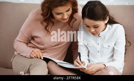 Kind schreibt auf Notizbuch in der Nähe von Mutter, die bei den Hausaufgaben hilft Stockfoto