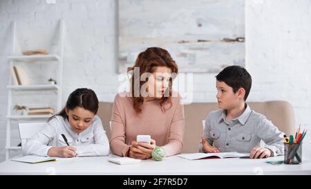 Mutter mit Smartphone sitzt neben Kindern, die Hausaufgaben machen Stockfoto