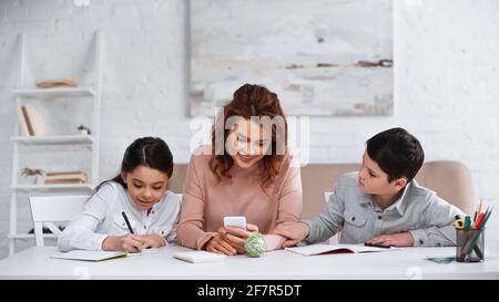 Lächelnde Mutter mit Smartphone macht Hausaufgaben mit Kindern im Leben Zimmer Stockfoto