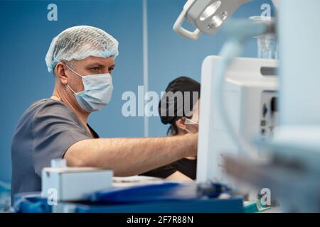 Anästhesist, der während der Herzoperation die lebenswichtigen Funktionen des Körpers verfolgt. Chirurg, der während der Operation einen medizinischen Monitor ansieht. Doktor Checki Stockfoto
