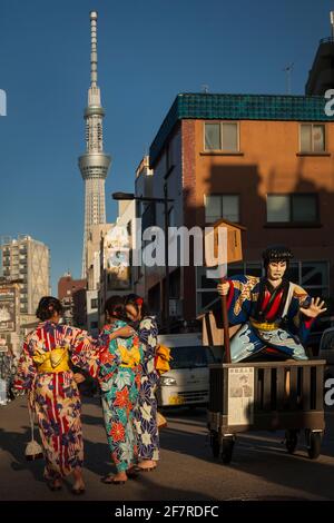 Drei junge Frauen, die traditionelle Yukatas tragen, kommen an einer japanischen Werbefigur mit Tokyo Sky Tree im Hintergrund vorbei, Asakusa, Tokio Stockfoto