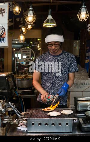 Vertikale Nahaufnahme eines jungen Kochs, der eine Jakobsmuschel in einem Verkaufsstand für Zwischenhändler im Bereich Meeresfrüchte auf dem Tsukiji Market, Tsukiji, Tokio, Japan, röstet Stockfoto