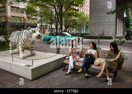 Horizontale Ansicht von drei jungen Frauen, die sich zum Mittagessen auf einer Marunouchi Naka-dori-Bank von einer Tiger-Skulptur entspannen, Ginza, Tokio, Japan Stockfoto