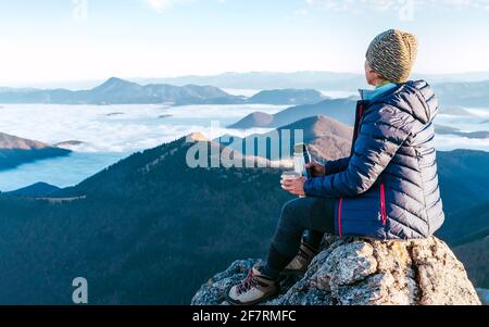 Junge Wandererin, die auf der Berggipfel-Klippe sitzt und die Aussicht auf das mit Wolken bedeckte Bergtal genießt. Erfolgreiches Gipfelkletterkonzept ima Stockfoto