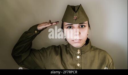 Porträt einer jungen Frau in Uniform auf dem Hintergrund der grauen Wand. Frau in sowjetischer Militäruniform während des Zweiten Weltkriegs grüßt Stockfoto