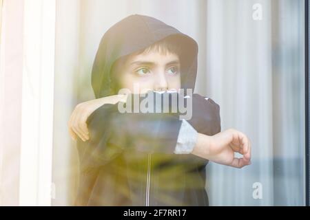 Trauriges Kind schaut aus dem Fenster glass pandemische Zeit Stockfoto