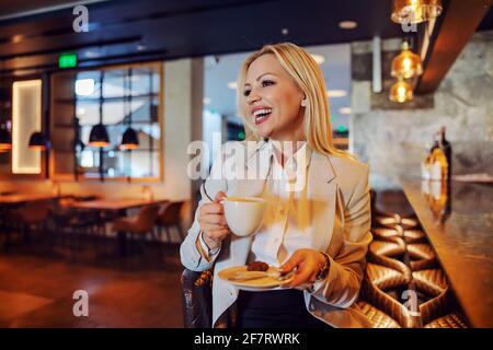 Selbstbewusste und lächelnde blonde Frau, elegant gekleidet, sitzt an der Bar im Hotelrestaurant und hat ihren Kaffee getrunken. Business Lifestyle, Taking a bre Stockfoto
