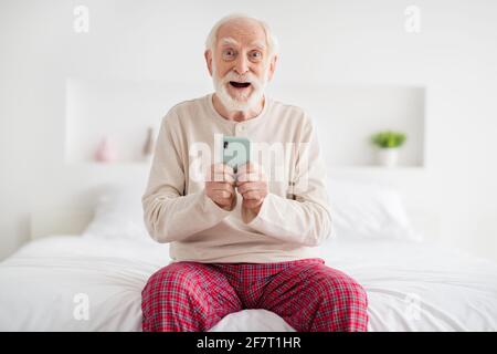 Foto von beeindruckt lustig reifen Mann Nachtwäsche sitzend Bett tippen Modernes Gerät im Innenbereich Stockfoto