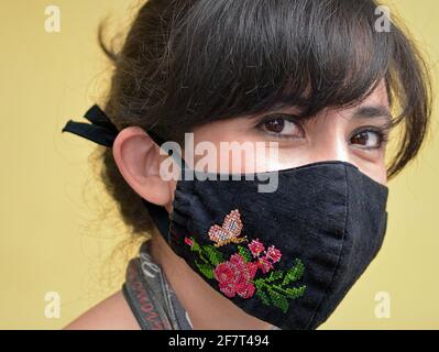 Die junge mexikanische Frau mit schönen dunklen Augen trägt während der Coronavirus-Pandemie eine kreuzgenähte, bestickte Stoffmaske mit Blumenmuster. Stockfoto