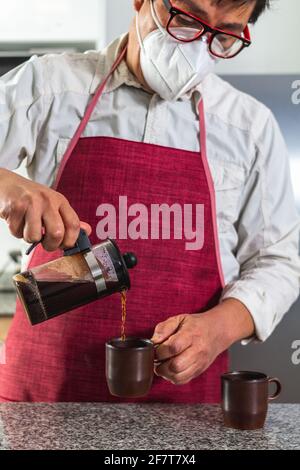Mann in roter Schürze und Gesichtsmaske Gießen zwei Tassen Von Kaffee in einer französischen Presse in einem Café Stockfoto