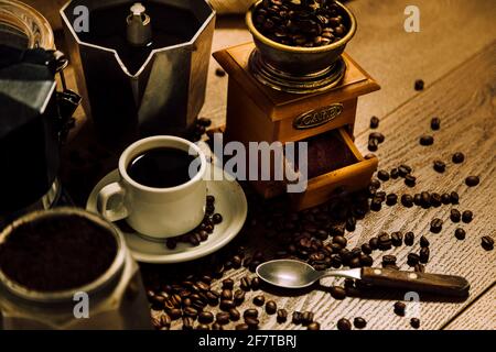 Kaffee. Eine weiße Tasse mit schwarzem Kaffee, italienische Kaffeemaschine Moka, eine Mühle und Getreide über einem Holztisch. Rustikaler Vintage-Stil. Stockfoto