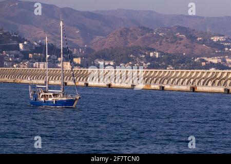 Ein blaues Segelboot, das den Hafen von Malaga vor einem verlässt Dock mit der Stadt im Hintergrund Stockfoto