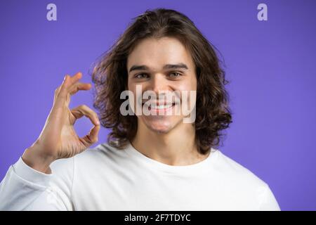 Gutaussehender Mann zeigt okay, ok Geste. Glücklicher Kerl auf violettem Hintergrund. Gewinner. Erfolg. Positive männliche Modell lächelt zur Kamera. Körpersprache. Stockfoto
