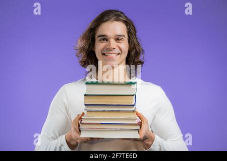 Student hält Stapel von Universitätsbüchern aus der Bibliothek auf violettem Hintergrund im Studio. Der Mann lächelt, er ist glücklich, seinen Abschluss zu machen. Stockfoto