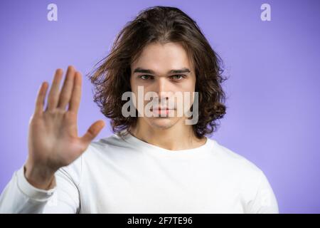 Porträt eines ernsten Mannes, der eine ablehnende Geste durch ein Schild mit der Stopphandhand zeigt. Kerl mit langer Frisur isoliert auf violettem Hintergrund. Stockfoto