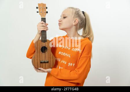 Glückliches Mädchen mit Ukulele in ihren Händen sieht seitwärts aus und lächelt, trägt gelbe Freizeitkleidung. Lächelnde Musikerin spielt Ukulele. Isoliert Stockfoto