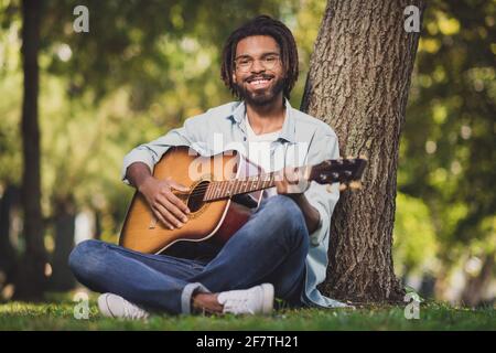 Portrait von attraktiven talentierten fröhlichen Kerl auf Gras sitzen spielen Gitarre verbringen Wochenende sonnigen Tag draußen Stockfoto