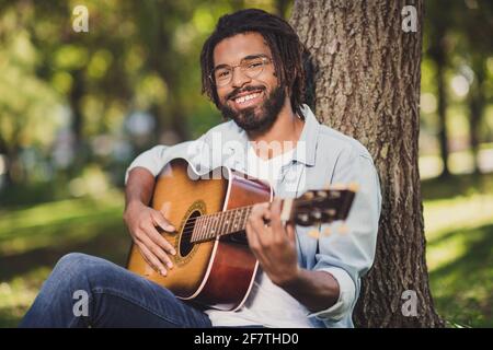 Foto-Porträt eines Mannes in einer Brille lächelnd sitzt in grün park unter Baum spielen Gitarre in lässiger Kleidung Stockfoto