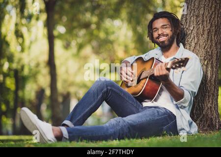 Portrait von attraktiven fröhlichen Kerl auf grünem Gras sitzen spielen Gitarre singen romantischen Hit sonnigen Tag draußen Stockfoto