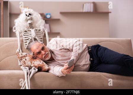 Alter Mann sitzt auf dem Sofa mit dem weiblichen Skelett In unsterbst Liebe Konzept Stockfoto