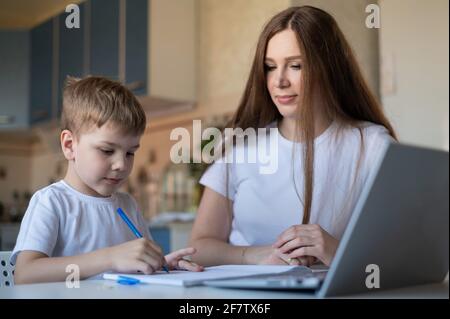 Kaukasische Frau hilft ihrem Sohn, Unterricht zu lernen. Die fürsorgliche Mutter prüft die Hausaufgaben des Kindes. Untersuchung zu Hause während der Quarantäne. Stockfoto