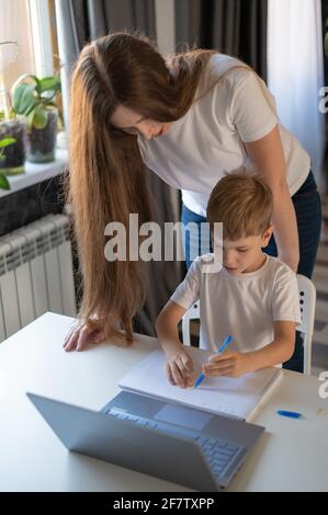 Kaukasische Frau hilft ihrem Sohn, Unterricht zu lernen. Die fürsorgliche Mutter prüft die Hausaufgaben des Kindes. Untersuchung zu Hause während der Quarantäne. Stockfoto