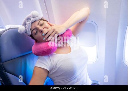 Die Frau gähnt, während sie mit einer Maske und einem Kissen zum Schlafen im Flugzeug sitzt. Stockfoto