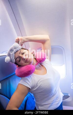 Die Frau gähnt, während sie mit einer Maske und einem Kissen zum Schlafen im Flugzeug sitzt. Stockfoto