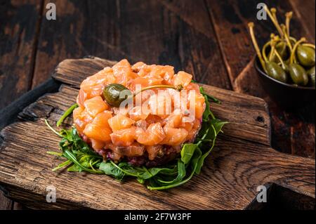 Lachstartar oder Tatar mit roten Zwiebeln, Avocado, Rucola und Kapern. Dunkler Holzhintergrund. Draufsicht Stockfoto