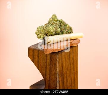 Ein verbranntes, handgewalztes Marihuana Joint gleicht auf rotem Sandstein mit zwei großen Marihuana-Knospen aus. Auf einem Holzständer gegen ein pfirsichfarbenes b geschossen Stockfoto