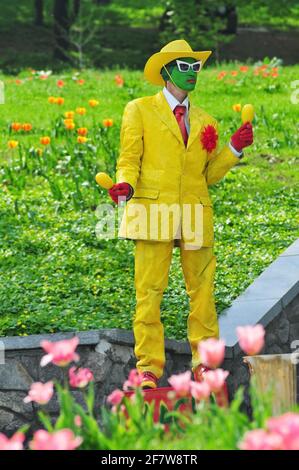 Junger Mann mit grün gemaltem Gesicht in Lederanzug gelb Und Hut zeigt eine Statue im Park Stockfoto