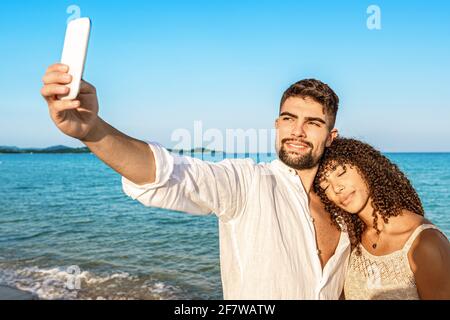 Ein paar junge Liebhaber machen ein Selfie im Urlaub am Meer bei Sonnenuntergang oder Sonnenaufgang mit Boho Sommerkleidung. Afroamerikanisches Model verliebt lehnt sich an sie, er Stockfoto