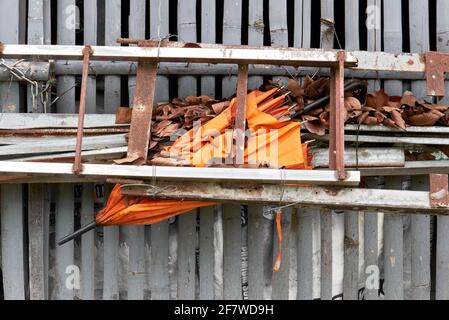 Nahaufnahme eines alten orangefarbenen gefalteten Schirms, der hinter einer Leiter zusammengedrückt und mit vielen alten Blättern an einem Gebäudezaun, Asien, bedeckt wurde Stockfoto