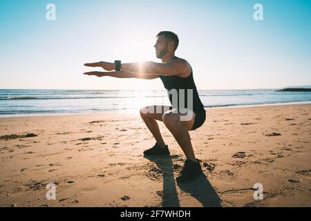 Mann, der während des Sonnenuntergangs hockt oder am Strand trainiert. Sport und Gesundheit Stockfoto