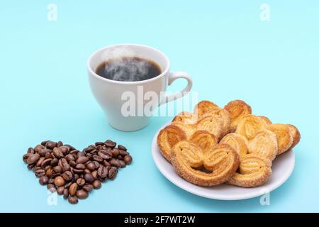 Dampfende Kaffeetasse, herzförmige Kekse, herzförmige Kaffeebohnen auf blauem Hintergrund. Frühstück am Morgen. Frühstück für die Liebsten. Stockfoto