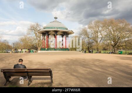 Ein junger Mann sitzt auf einer Parkbank neben Clapham Common Bandstand, Windmill Drive, Clapham Common, Clapham, SW4, London, England, Großbritannien Stockfoto