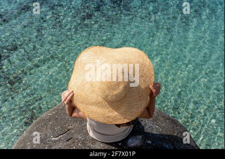 Blick von oben unkenntlich Frau trägt Strohhut auf einem Felsen sitzen Blick auf das schöne durchscheinende Meerwasser. Stockfoto