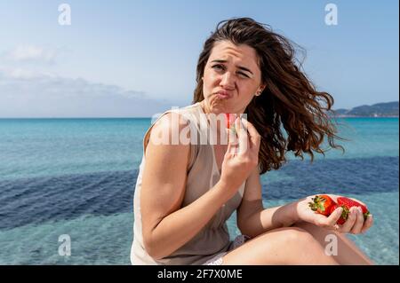 Junge Frau am Strand mit wunderschönem Meer im Hintergrund, die mit einem lustigen Gesicht auf die Kamera schaut, während sie eine Erdbeere isst. Stockfoto