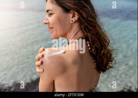 Rückseite einer jungen Frau, die Sonnencreme auf ihre Schulter applyte. Frau im Bikini am Strand. Stockfoto
