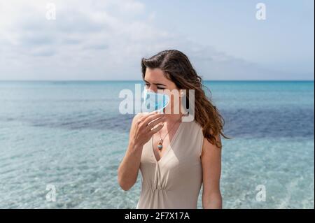 Porträt einer jungen Frau, die gegen blaues klares Wasser auf dem Meer steht. Weibchen nimmt eine chirurgische Schutzmaske ab Stockfoto