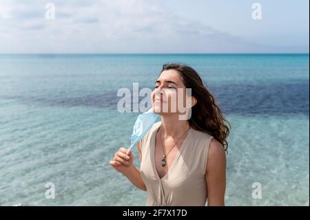 Porträt einer jungen lächelnden Frau, die eine chirurgische Schutzmaske auszieht, um am Strand reine frische Luft zu atmen. Schönes blaues klares Wasser auf der Rückseite Stockfoto