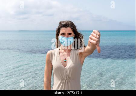 Porträt einer jungen Frau mit einer schützenden Gesichtsmaske, die den Daumen nach unten zeigt. Schöner Hintergrund mit blauem klarem Wasser. Stockfoto