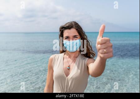 Porträt einer jungen Frau mit einer schützenden Gesichtsmaske, die den Daumen nach oben zeigt. Schöner Hintergrund mit blauem klarem Wasser Stockfoto