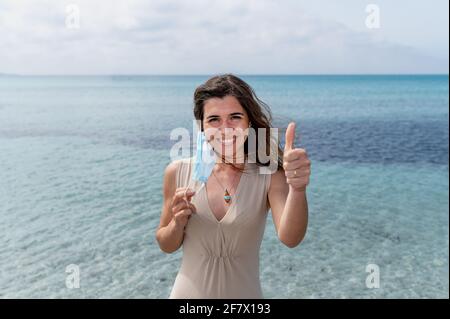 Horizontales Banner oder Kopfzeile Porträt einer jungen Frau, die eine schützende Gesichtsmaske auszieht und den Daumen nach oben zeigt. Schöner Hintergrund mit blauem klarem Wasser Stockfoto