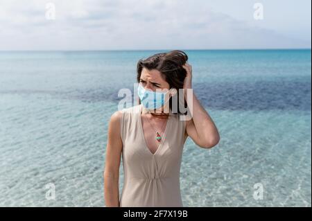 Eine junge Frau, die eine schützende Gesichtsmaske mit einem besorgten Gesicht trägt, hält ihre Hand hinter dem Kopf. Meer auf dem Hintergrund. Stockfoto