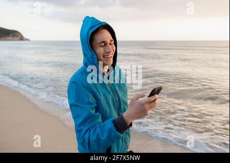 Seitenansicht junger Gelegenheitsmann, der am Strand Smartphone benutzt, chattet und SMS schreibt. Stockfoto