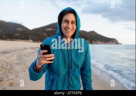 Junge kausale Mann Porträt hält Smartphone und Blick auf die Kamera mit selbstbewussten Smiley Gesicht am Strand. Stockfoto