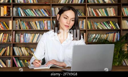Attraktive junge Dame in stilvoller weißer Bluse schreibt in Papier Notebook auf Videocall auf einem modernen Laptop am Tisch gegen Racks Von Büchern Stockfoto
