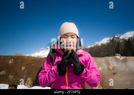 Frostiger Winterurlaub - jung glücklich und schön asiatisch koreanisch Frau auf der Bank am gefrorenen See und an den Schneebergen trinkt Kaffee oder heiße Schokolade en Stockfoto