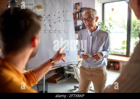 Eine ältere Chefin hört sich die Frage ihres männlichen Kollegen genau an, während sie in einer Arbeitsatmosphäre am Arbeitsplatz eine Präsentation hält. Bus Stockfoto